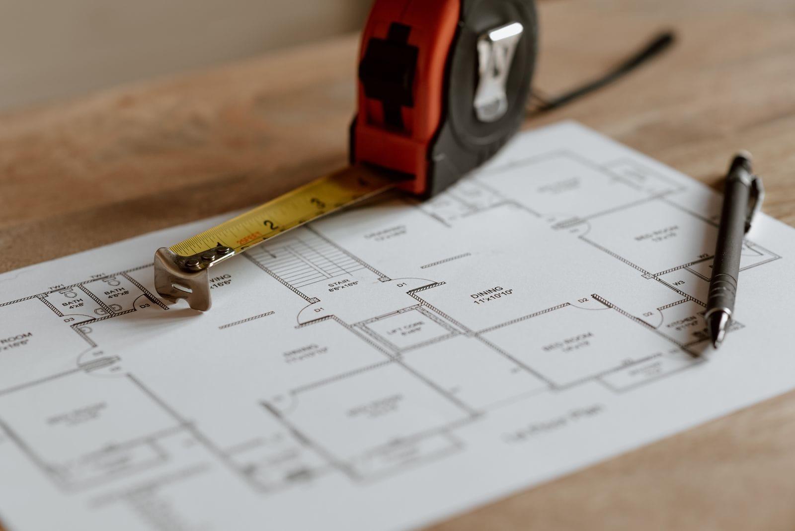 Architectural floor plan on a wooden desk with a tape measure and a pen